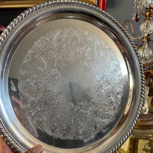 May include: A round silver-toned serving tray featuring a polished center and an intricate, embossed floral scrollwork pattern around the inner rim. The tray is finished with a thick, twisted rope-style border along the outer edge.