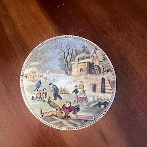 May include: A vintage ceramic lid featuring a winter scene of people ice skating. The lid is white with a blue and white border. The scene is painted in muted colours and includes a small house with a ladder, trees, and a figure falling on the ice. The text "DANGEROUS" is written on the ice.