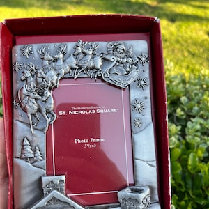 May include: A silver-colored photo frame with a Christmas theme. The frame features Santa Claus in a sleigh pulled by reindeer, snowflakes, and a house with a chimney. The text "Photo Frame 3 1/2x5" is visible.