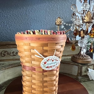May include: A woven basket with a light brown hue, featuring a decorative pink ribbon and a tag that reads "Snapdragon". The basket has a flared shape and is set on a dark wooden surface. The top edge is lined with a patterned fabric.