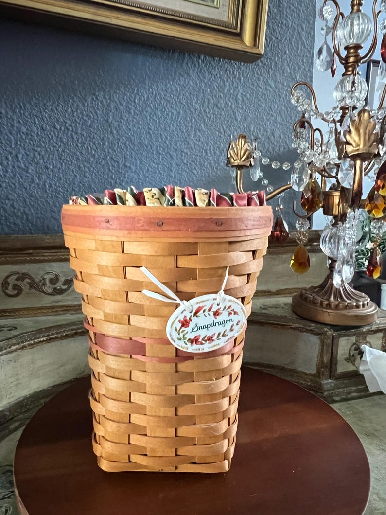 May include: A woven basket with a light brown hue, featuring a decorative pink ribbon and a tag that reads "Snapdragon". The basket has a flared shape and is set on a dark wooden surface. The top edge is lined with a patterned fabric.