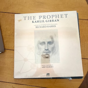 Puede incluir: Portada de álbum de vinilo con una ilustración en blanco y negro de un rostro humano y el título "El Profeta" de Kahlil Gibran. El álbum presenta una interpretación musical de Richard Harris con música compuesta por Arif Mardin.