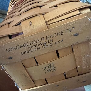 May include: A handwoven Longaberger basket made in Dresden, Ohio, USA. The basket is constructed from woven light brown wood strips. The text "LONGABERGER BASKETS HANDWOVEN DRESDEN, OHIO USA" is printed on the side. The basket has a handle and a lid.
