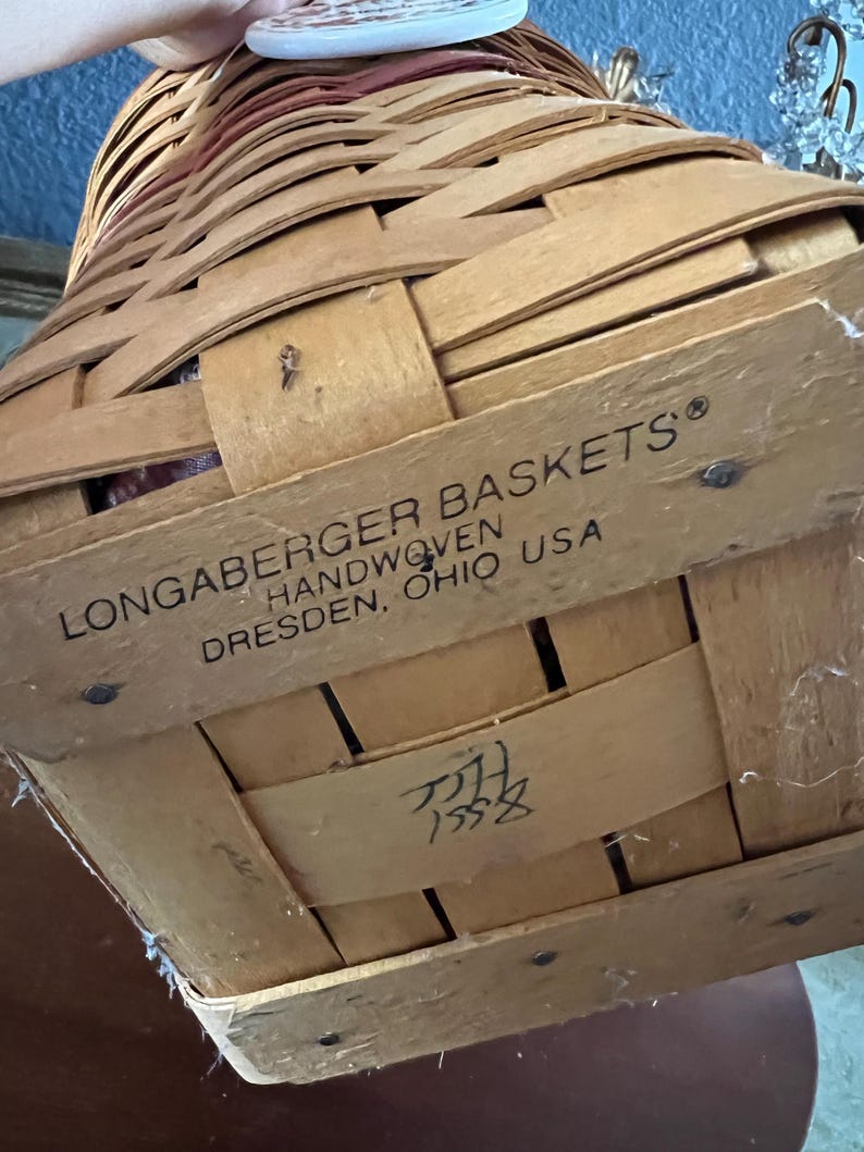May include: A handwoven Longaberger basket made in Dresden, Ohio, USA. The basket is constructed from woven light brown wood strips. The text "LONGABERGER BASKETS HANDWOVEN DRESDEN, OHIO USA" is printed on the side. The basket has a handle and a lid.