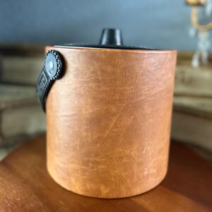 May include: A brown leather ice bucket with a black handle and lid. The bucket has a cylindrical shape and a textured surface. The handle is attached to the side with a decorative metal piece.