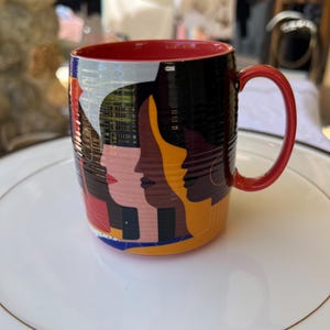 K&ouml;nnte beinhalten: Eine Keramik-Kaffeetasse mit rotem Innenraum und Henkel. Das &Auml;u&szlig;ere zeigt ein Design von stilisierten Gesichtern in verschiedenen Hautt&ouml;nen vor schwarzem Hintergrund. Die Tasse steht auf einem wei&szlig;en Teller mit Goldrand.