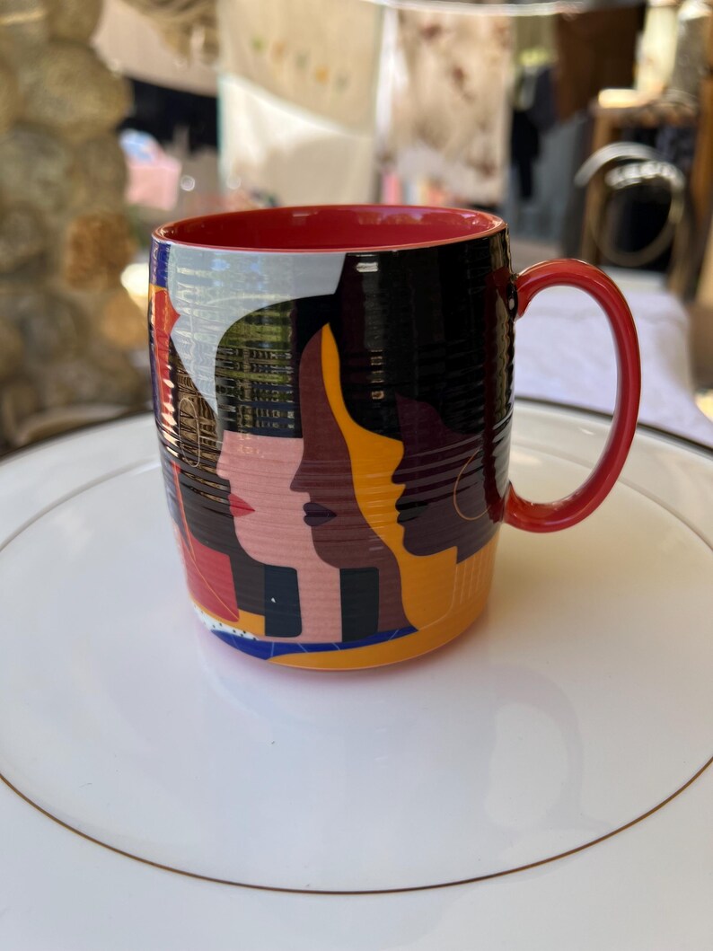K&ouml;nnte beinhalten: Eine Keramik-Kaffeetasse mit rotem Innenraum und Henkel. Das &Auml;u&szlig;ere zeigt ein Design von stilisierten Gesichtern in verschiedenen Hautt&ouml;nen vor schwarzem Hintergrund. Die Tasse steht auf einem wei&szlig;en Teller mit Goldrand.