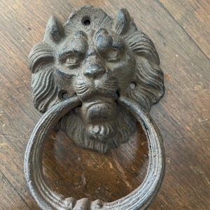 May include: A cast iron lion head door knocker with a ring handle. The knocker is dark brown and has a distressed finish.