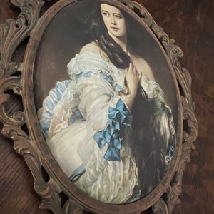 Puede incluir: Un retrato ovalado de una mujer con un vestido blanco con detalles azules, enmarcado en un marco de metal marr&oacute;n oscuro adornado.