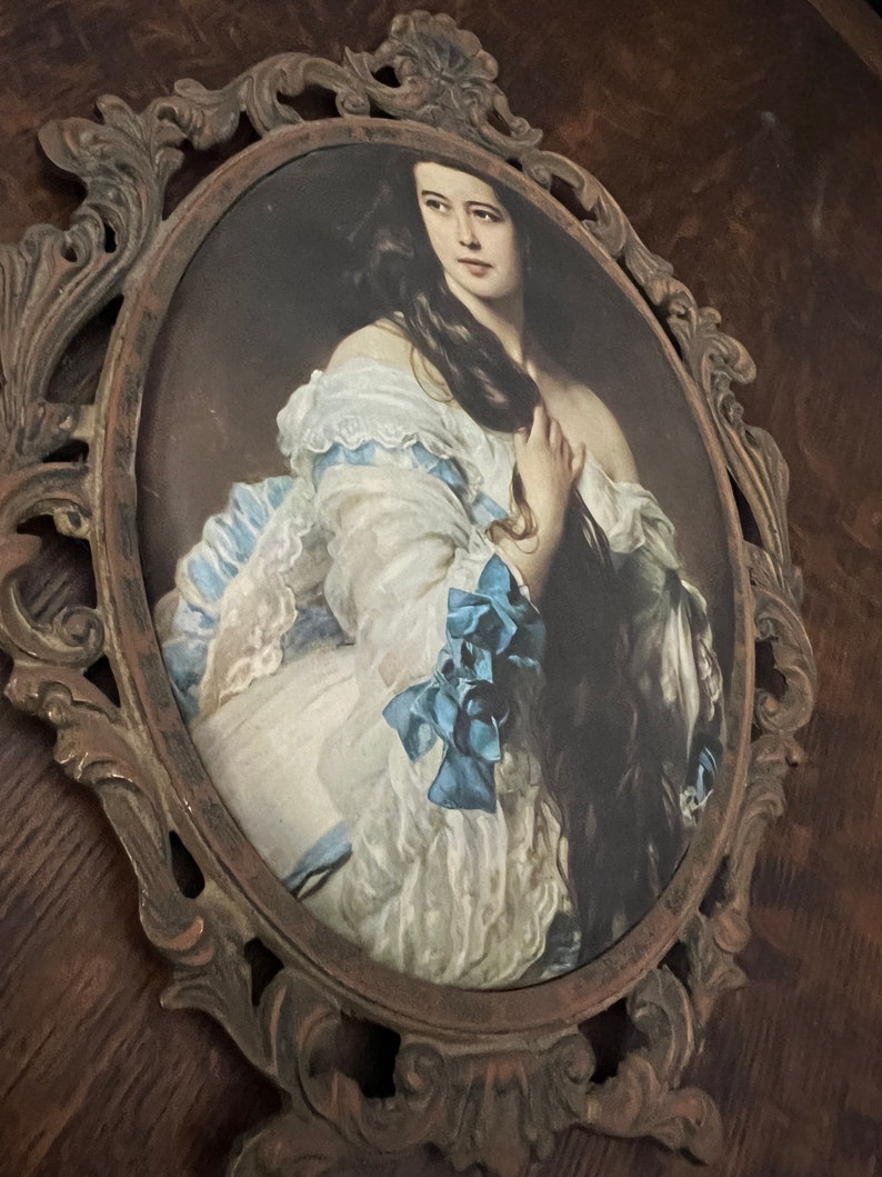 Puede incluir: Un retrato ovalado de una mujer con un vestido blanco con detalles azules, enmarcado en un marco de metal marr&oacute;n oscuro adornado.
