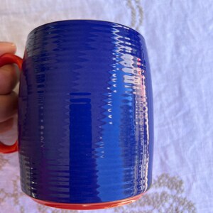 K&ouml;nnte beinhalten: Eine blaue Keramiktasse mit strukturierter Oberfl&auml;che und rotem Henkel und Boden. Die Tasse ist zylindrisch geformt und scheint leer zu sein. Die Tasse steht auf einer wei&szlig;en Oberfl&auml;che.