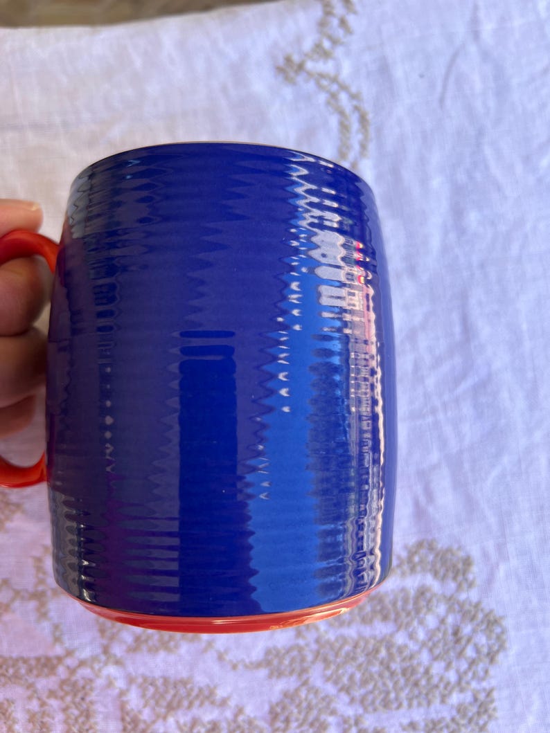 K&ouml;nnte beinhalten: Eine blaue Keramiktasse mit strukturierter Oberfl&auml;che und rotem Henkel und Boden. Die Tasse ist zylindrisch geformt und scheint leer zu sein. Die Tasse steht auf einer wei&szlig;en Oberfl&auml;che.