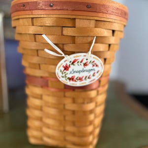 May include: A woven basket with a rectangular shape, featuring a light brown color with reddish-brown accents. A decorative tag with the word "Snapdragon" and floral designs hangs from the basket. The basket is likely used for storage or display.