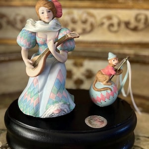 May include: A porcelain musical figurine set featuring a woman in a harlequin costume playing a lute and a monkey on a ball. The set is on a black wooden base with a silver plaque.