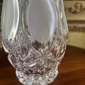 May include: A clear, cut-glass perfume bottle with a diamond pattern and oval frosted panels. The bottle has a rounded base and a flared, faceted top. The glass reflects light, creating a sparkling effect.