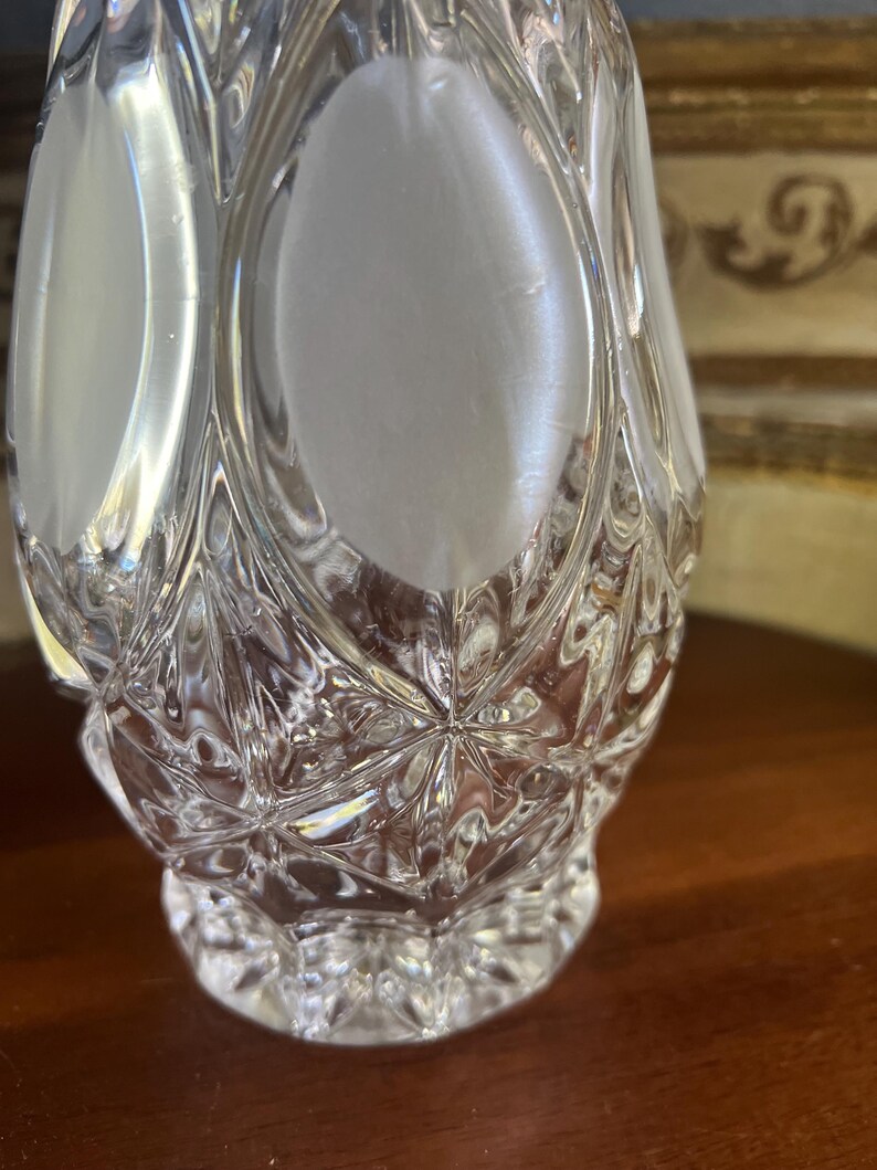 May include: A clear, cut-glass perfume bottle with a diamond pattern and oval frosted panels. The bottle has a rounded base and a flared, faceted top. The glass reflects light, creating a sparkling effect.