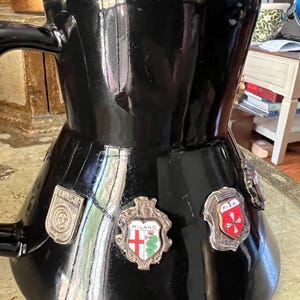 May include: A black ceramic coffee pot with a handle and a wide base. The pot has several metal badges with city names and emblems, including "Milano" and "Pisa". The pot has a glossy finish and is sitting on a light-colored surface.