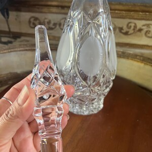 May include: A clear crystal decanter with a matching stopper. The decanter has a diamond pattern with frosted oval accents. The stopper is held in a hand and has a diamond pattern. The background is a blurred image of a gold-framed mirror.