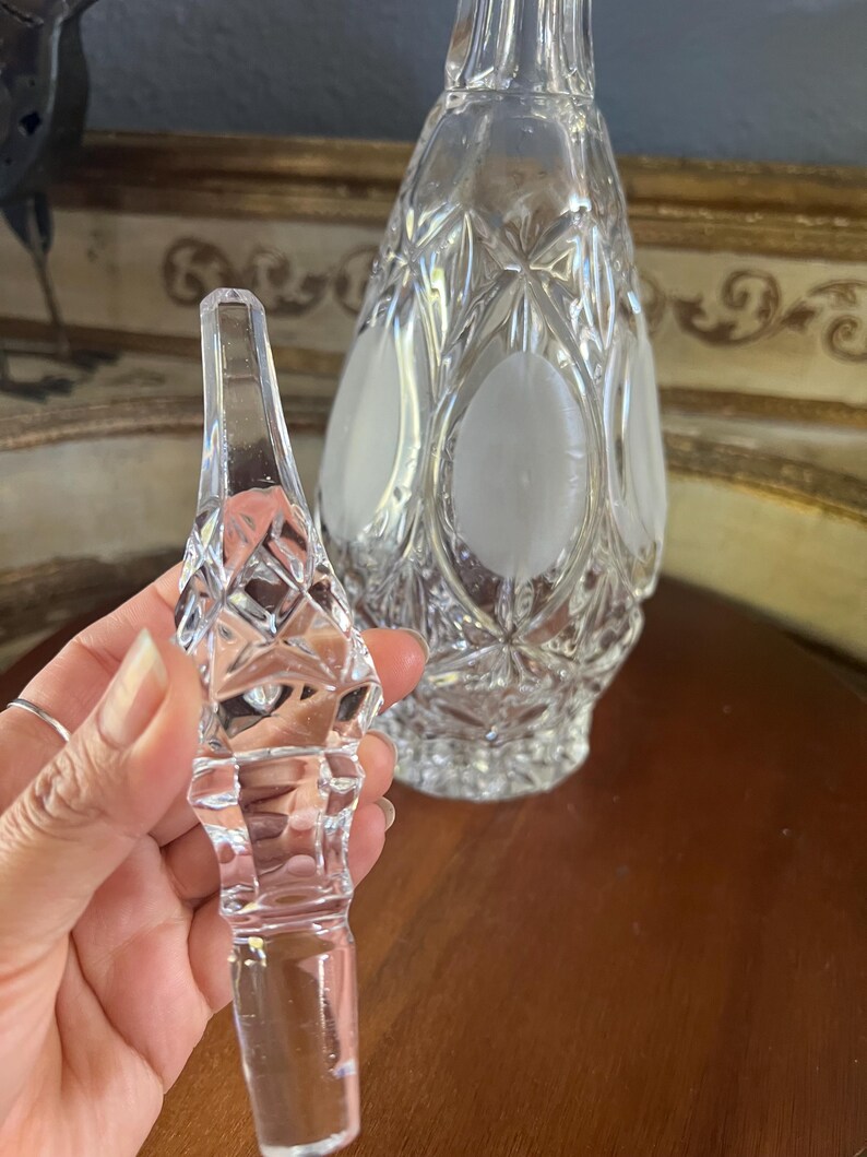 May include: A clear crystal decanter with a matching stopper. The decanter has a diamond pattern with frosted oval accents. The stopper is held in a hand and has a diamond pattern. The background is a blurred image of a gold-framed mirror.