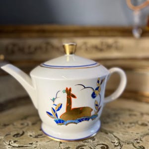 May include: White porcelain teapot with a gold trim and a deer design in gold, blue, and orange. The teapot has a lid with a gold knob.