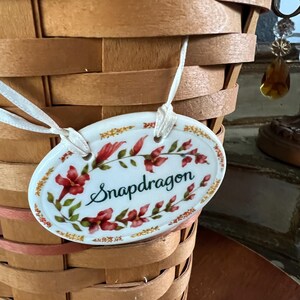 May include: A woven brown basket with a white oval tag. The tag is decorated with red flowers and green leaves, with the word "Snapdragon" in cursive script. The tag is attached to the basket with a white ribbon.