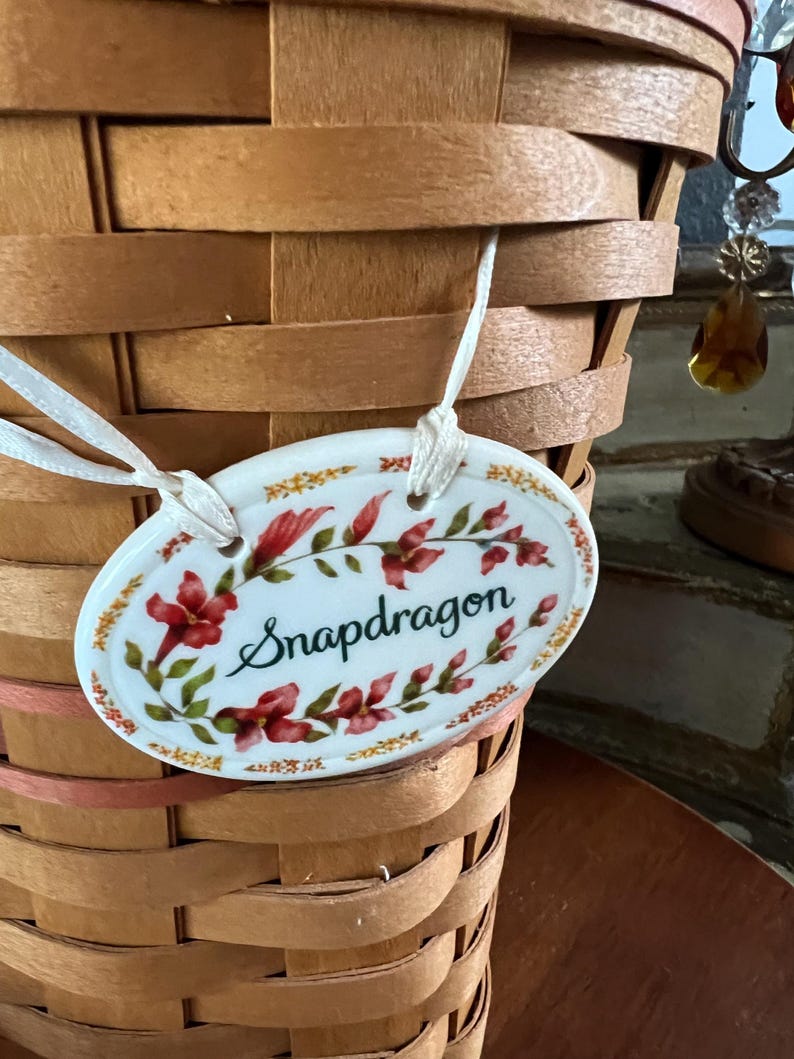 May include: A woven brown basket with a white oval tag. The tag is decorated with red flowers and green leaves, with the word "Snapdragon" in cursive script. The tag is attached to the basket with a white ribbon.