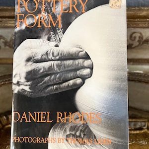 May include: A book titled "Pottery Form" by Daniel Rhodes with photographs by Thomas  Luken. The cover features a black and white photograph of a person's hands shaping a clay pot.