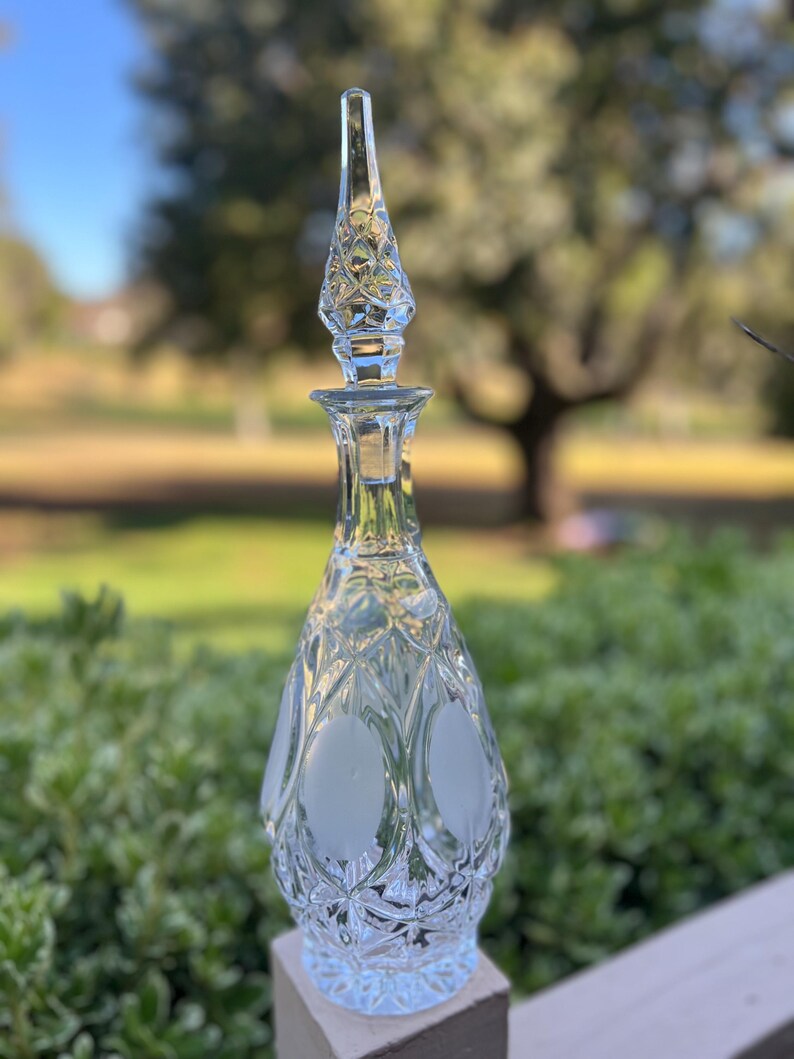 May include: A clear glass decanter with a diamond pattern and a frosted oval design. The stopper is tall and pointed, with a diamond pattern. The decanter is likely used for serving wine or other beverages.