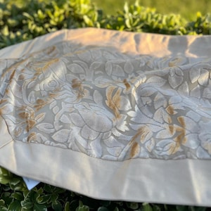 May include: A beige decorative table runner with a floral pattern in gold and white. The runner has a solid beige border and is made of a soft, textured fabric. The runner is placed on a green surface.