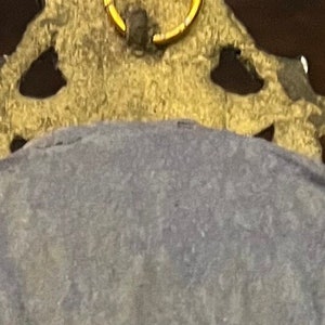 Puede incluir: Un marco de metal dorado con un dise&ntilde;o ornamentado. El marco tiene un anillo en la parte superior para colgar. La parte posterior del marco est&aacute; cubierta con una tela azul oscuro.