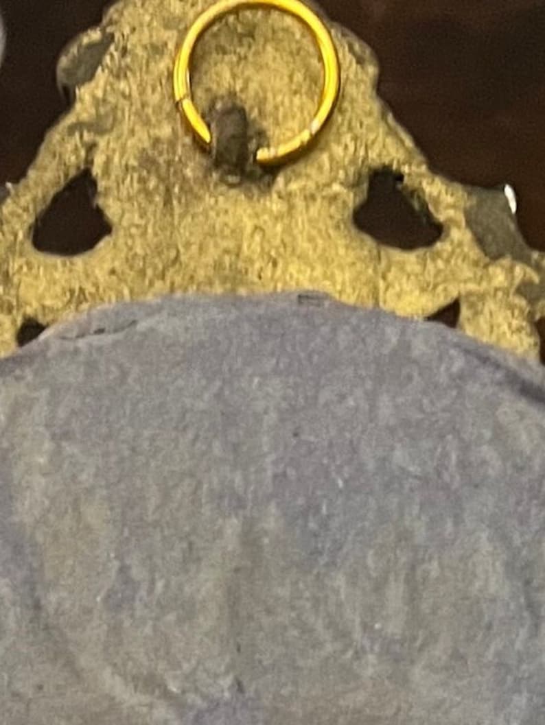Puede incluir: Un marco de metal dorado con un dise&ntilde;o ornamentado. El marco tiene un anillo en la parte superior para colgar. La parte posterior del marco est&aacute; cubierta con una tela azul oscuro.