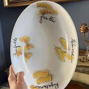 May include: An oval white ceramic serving bowl featuring hand-painted illustrations of various pasta shapes including fusilli, farfalle, penne, and rigatoni. Each pasta type is labeled with its corresponding name in black cursive script lettering.