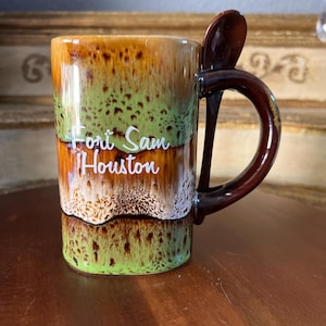 May include: A ceramic mug with a brown handle and a spoon attached. The mug has a gradient design of brown, green, and white. The words "Fort Sam Houston" are printed on the mug.