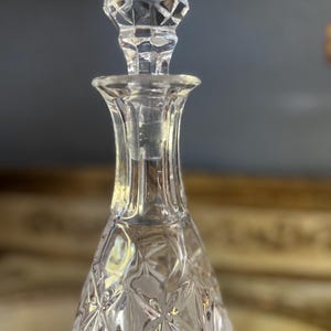 May include: A clear glass decanter with a diamond-cut pattern and a faceted stopper. The stopper is a geometric shape, and the decanter has a flared neck. The design is classic and elegant, suitable for serving beverages.