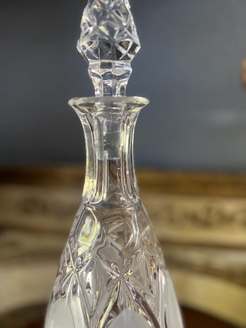 May include: A clear glass decanter with a diamond-cut pattern and a faceted stopper. The stopper is a geometric shape, and the decanter has a flared neck. The design is classic and elegant, suitable for serving beverages.