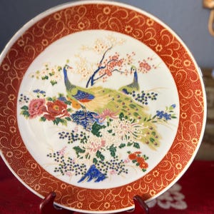 May include: A decorative ceramic plate featuring a peacock design. The plate has a white center with two peacocks, flowers, and a tree. The rim is a reddish-orange color with a detailed pattern. The plate is displayed on a wooden stand.