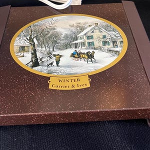 May include: A brown metal electric warming tray with a gold oval design featuring a winter scene with a horse-drawn carriage and a house. The tray has the text "WINTER" and "Currier & Ives" in gold lettering.