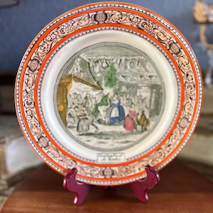 May include: A decorative ceramic plate with a detailed illustration of a gathering scene. The plate features a wide orange border with ornate floral and scroll designs. The central image depicts people in a room, with text at the bottom. The plate is displayed on a red stand.