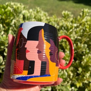 K&ouml;nnte beinhalten: Eine Keramiktasse mit einem farbenfrohen abstrakten Design von vier verschiedenen Gesichtern im Profil. Die Tasse hat einen roten Henkel und eine lebendige Farbpalette mit Rot, Schwarz, Gelb und Blau. Die Tasse wird vor einem Hintergrund aus gr&uuml;nem Laub gehalten.