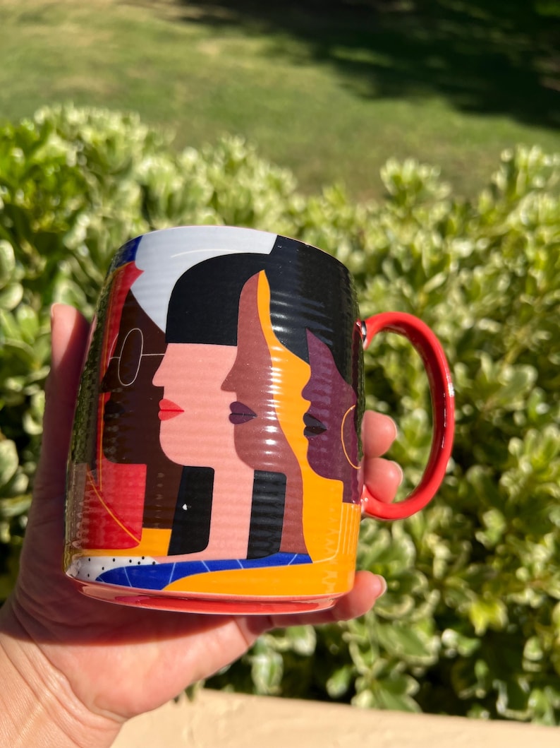 K&ouml;nnte beinhalten: Eine Keramiktasse mit einem farbenfrohen abstrakten Design von vier verschiedenen Gesichtern im Profil. Die Tasse hat einen roten Henkel und eine lebendige Farbpalette mit Rot, Schwarz, Gelb und Blau. Die Tasse wird vor einem Hintergrund aus gr&uuml;nem Laub gehalten.