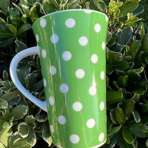May include: A green ceramic pitcher with white polka dots. The pitcher has a white handle and a wide opening. The background is a lush green bush. This pitcher is perfect for serving beverages.