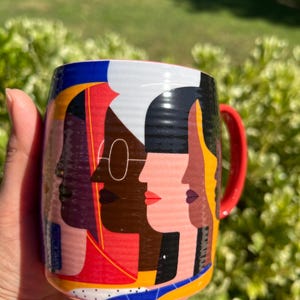K&ouml;nnte beinhalten: Eine Keramiktasse mit einem lebendigen Design, das stilisierte Gesichter in verschiedenen Hautt&ouml;nen zeigt. Die Tasse hat einen roten Henkel und ein farbenfrohes geometrisches Muster. Der Hintergrund ist unscharf, was auf eine Umgebung im Freien hindeutet.