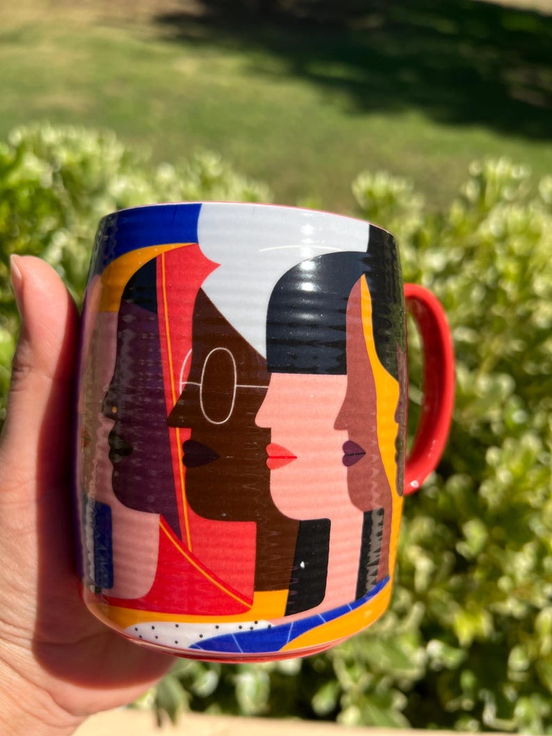 K&ouml;nnte beinhalten: Eine Keramiktasse mit einem lebendigen Design, das stilisierte Gesichter in verschiedenen Hautt&ouml;nen zeigt. Die Tasse hat einen roten Henkel und ein farbenfrohes geometrisches Muster. Der Hintergrund ist unscharf, was auf eine Umgebung im Freien hindeutet.