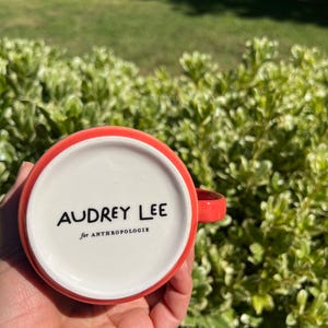 K&ouml;nnte beinhalten: Eine rot-wei&szlig;e Keramiktasse mit dem Text "AUDREY LEE for ANTHROPOLOGIE" auf der Unterseite. Die Tasse hat einen roten Henkel und Rand. Die Tasse wird in der Hand einer Person gehalten.