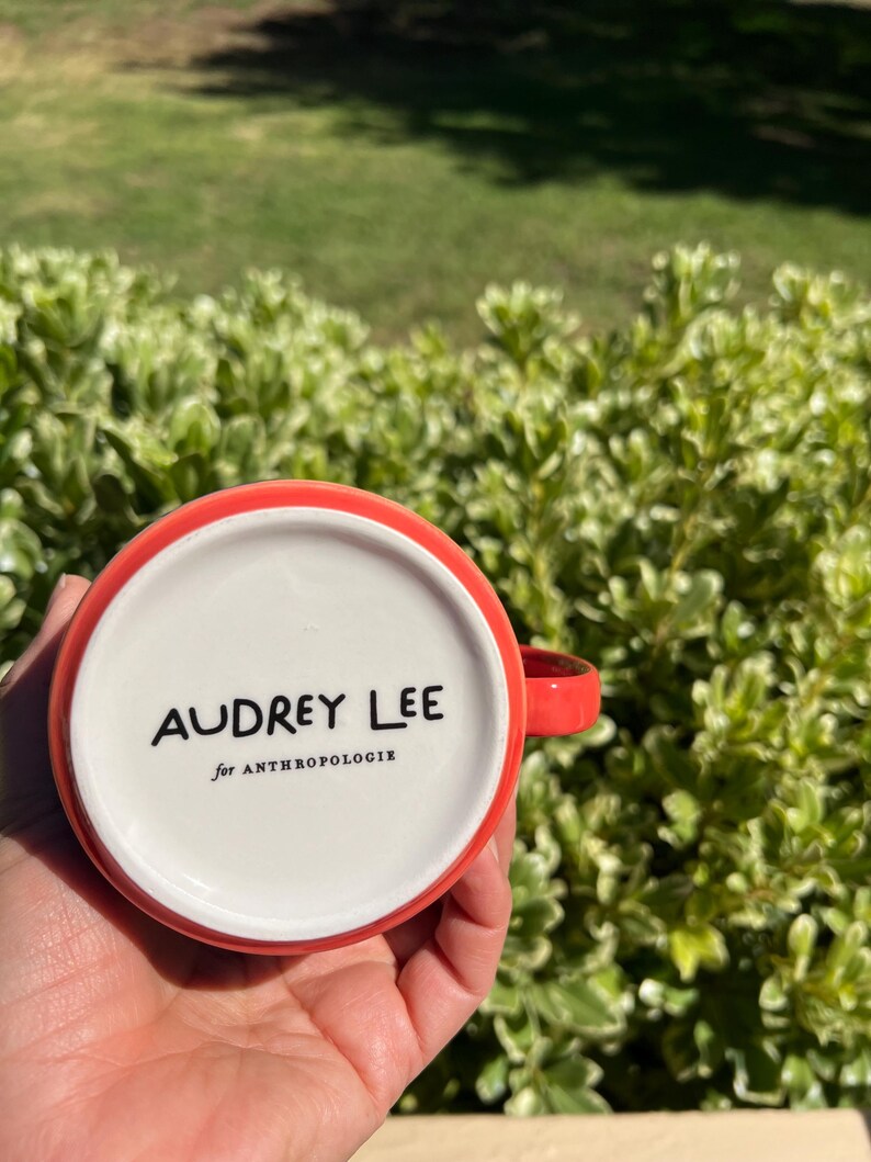 K&ouml;nnte beinhalten: Eine rot-wei&szlig;e Keramiktasse mit dem Text "AUDREY LEE for ANTHROPOLOGIE" auf der Unterseite. Die Tasse hat einen roten Henkel und Rand. Die Tasse wird in der Hand einer Person gehalten.