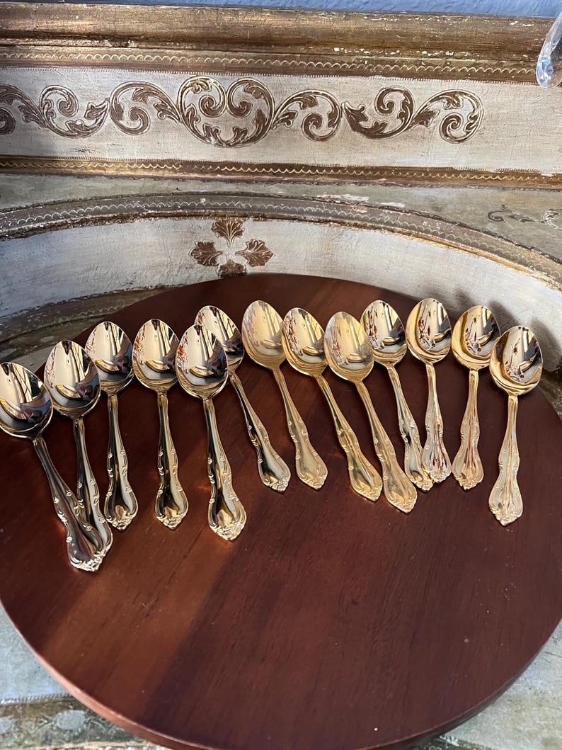 May include: A set of 12 gold-plated teaspoons with ornate handles. The spoons are arranged in a semi-circle on a wooden surface.