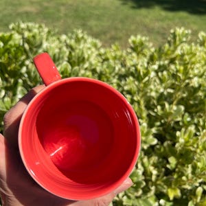 K&ouml;nnte beinhalten: Eine leuchtend rote Keramik-Tasse mit passendem Henkel. Das Innere der Tasse ist leuchtend rot, und die Au&szlig;enseite ist ein hellerer Rotton. Die Tasse wird vor einem Hintergrund aus gr&uuml;nem Laub gehalten.
