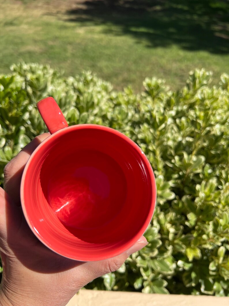 K&ouml;nnte beinhalten: Eine leuchtend rote Keramik-Tasse mit passendem Henkel. Das Innere der Tasse ist leuchtend rot, und die Au&szlig;enseite ist ein hellerer Rotton. Die Tasse wird vor einem Hintergrund aus gr&uuml;nem Laub gehalten.