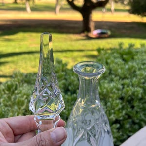 May include: A clear glass bottle and stopper set. The stopper has a diamond pattern and a pointed top. The bottle has a rounded shape with a narrow neck. The set is likely for decorative use.