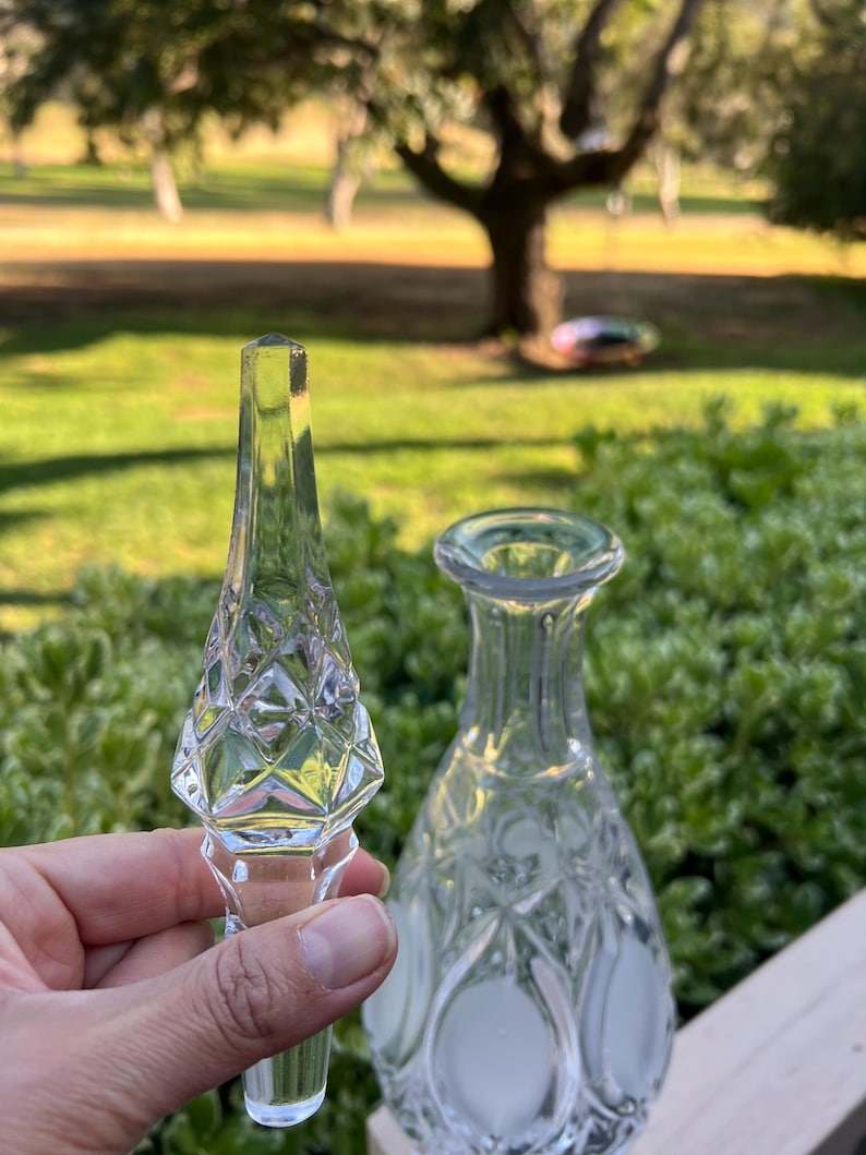 May include: A clear glass bottle and stopper set. The stopper has a diamond pattern and a pointed top. The bottle has a rounded shape with a narrow neck. The set is likely for decorative use.